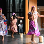 Women singing and dancing on a stage, holding mortar and pestle tools used to mash plantains.