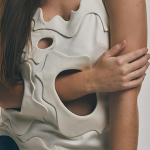 Image of a model in a white wool top with curvilinear tonal appliqués with smooth holes tailored into the shapes. The model is reaching her arms through the voids in the top