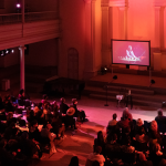 A large hall is lit in red. Guests are seated as an individual gives remarks at the front right of the hall, a sign language interpreter beside them. A screen in the center of the space features an individual onscreen who appears to be pictured in the midst of a musical performance.