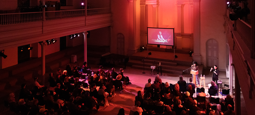 A large hall is lit in red. Guests are seated as an individual gives remarks at the front right of the hall, a sign language interpreter beside them. A screen in the center of the space features an individual onscreen who appears to be pictured in the midst of a musical performance.