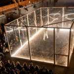 A large performance box lit with white light and clear walls/ceiling is placed in a large, brick-walled industrial space in Brooklyn. Two individuals/performers inside are being watched by onlooker on the outside.