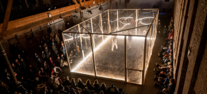 A large performance box lit with white light and clear walls/ceiling is placed in a large, brick-walled industrial space in Brooklyn. Two individuals/performers inside are being watched by onlooker on the outside.