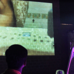 An individual looks up at a person's face projected on a large screen, purple lights casting a soft glow on the righthand side of their face
