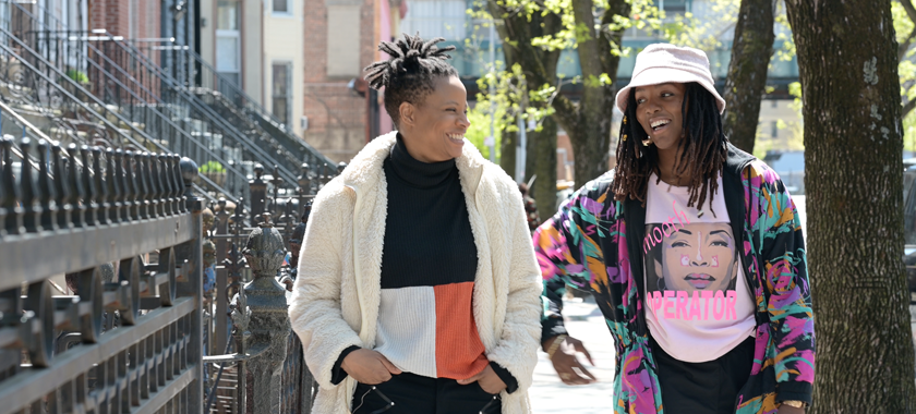 Two individuals in bright patterns appear to be laughing while walking down a sidewalk in a city setting.