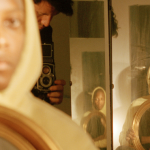 A portrait of Elliott Jerome Brown Jr. wearing a yellow hoodie in front of a mirror that reflects a trifold mirror that lies between he and the photographer, Matthew Morrocco.
