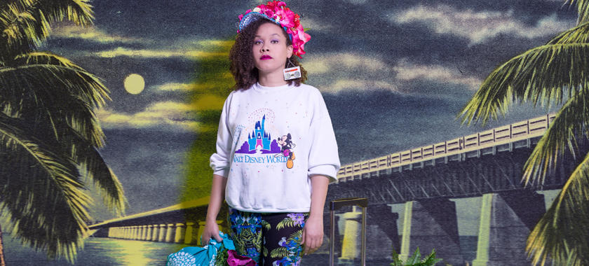 Self portrait of a Caribbean woman standing in front of a composited postcard landscape of palm trees and on top of the ocean dressed in tropical patterns and a Disney World sweatshirt, carrying luggage and tropical plants