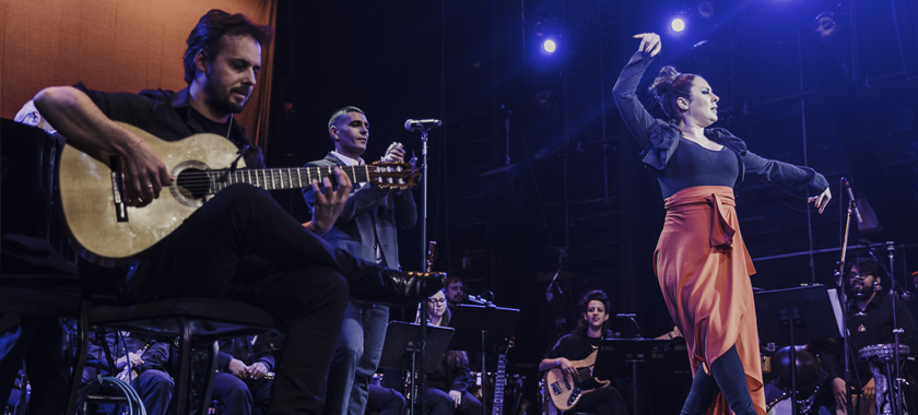 Sonia Olla dancing in a red and black dress with a guitarist in the foreground and an orchestra in the back.