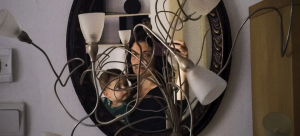 A self-portrait of Rachel Papo holding her son, reflected through a vertical oval mirror and obstructed by a lamp structure made up of a tangle of wires and lampshades.