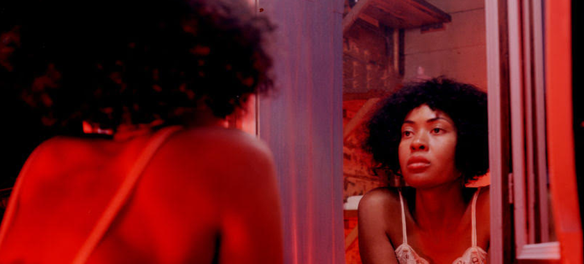 A woman in a nightgown looks intently into the mirror while resting her arms on the edge of the sink.