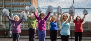 Six older women cheerleaders dressed colorfully with their silver pom-poms raised above their heads as they cheer from the rooftop of the 14th Street Y on a sunny day.