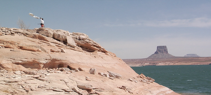 An individual is standing at the top of a rock formation, one arm raised with a white scarf flying in the wind behind them. There is a body of water below, and a butte and a mesa in the distance.