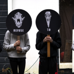 Four children stand in a row in front of a dark gray shed in the Pacific Northwest, holding circular protest signs that shield their faces from view. Printed in white on a black background, each sign illustrates a human or animal skull facing head-on, rendered in the style of a woodblock print. Underneath each skull is one word of hand-drawn text, which together compose the sentence “WE REFUSE TO DIE.”