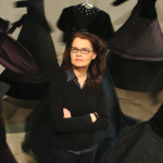 Alyson Pou with her installation"Where in Fury Takes Flight." She stands with her arms crossed wearing black, in between what appear to be black garments dynamically installed in a white-walled gallery space.