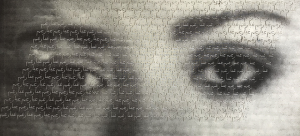 Photo litho print of large eyes gazing directly ahead at the viewer. There are laser-cut words on the eyes and eyebrows which are Persian repeated words and the area of the cut text makes up a parallelogram and the overall color scheme is different shades of gray.