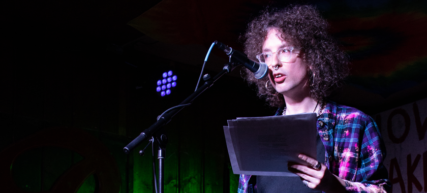 imogen, wearing bleach-purple flannel, black top & leather pants, gestures while reading onstage at Blue Moon.