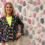 An individual stands smiling at the camera, pictured in front of an installation of pink, cream, and grey sculptures affixed to a white gallery wall