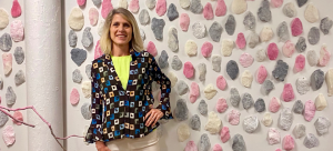 An individual stands smiling at the camera, pictured in front of an installation of pink, cream, and grey sculptures affixed to a white gallery wall