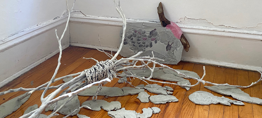 A grouping of white/grey twigs roped together, with grey ceramic fragments on a wooden floor against a slightly crumbling white wall