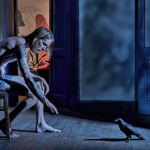 A half naked man sits in an old empty room facing a large balcony door. A nocturnal light illuminates him while he throws seeds on the floor to feed a black crow who has just walked into the space.
