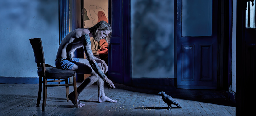 A half naked man sits in an old empty room facing a large balcony door. A nocturnal light illuminates him while he throws seeds on the floor to feed a black crow who has just walked into the space.