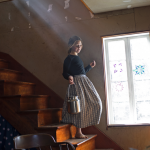 Barefoot, a young girl in a long skirt carries a metal container as she descends the staircase of her home, bathed in sunlight from an interior window. She is headed outdoors to collect maple tree sap on the family farm.