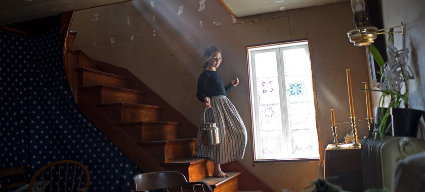 Barefoot, a young girl in a long skirt carries a metal container as she descends the staircase of her home, bathed in sunlight from an interior window. She is headed outdoors to collect maple tree sap on the family farm.