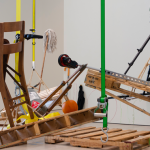 A group of ordinary objects is precariously arranged over three pallets, suspended on the border of collapse. Throughout the exhibition, the objects, initially leaning against the gallery walls, are activated and moved to be part of the composition in fragile balance. On the walls, colorful outline drawings record the objects' displacement.