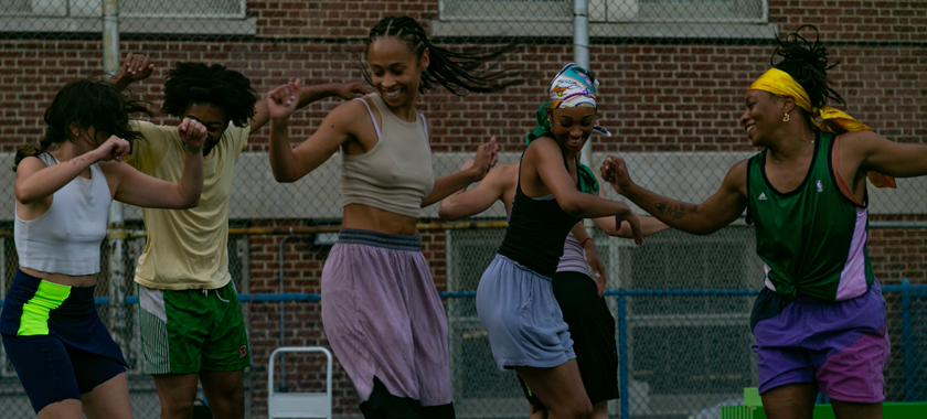 Six dancers wearing athletic clothing and sneakers lift their arms and twists their spines in the moments before jumping off the ground. On slightly different facings and ripples in their fabric of their clothes, the collective moves with soft smiles and subtle expressions of joy.