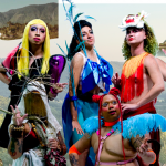 Five colorfully dressed ensemble members from a performance called "TERRITORY: The Island Remembers" pose in shimmering costumes, wigs, and make up. A few give fierce looks to the camera lens standing and kneeling in front of a background of a collage featuring mountains, lush green hills, trees, and other vegetation.