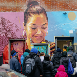 People line up outside a brick building with a colorful mural painted over some of the bricks