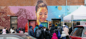 People line up outside a brick building with a colorful mural painted over some of the bricks