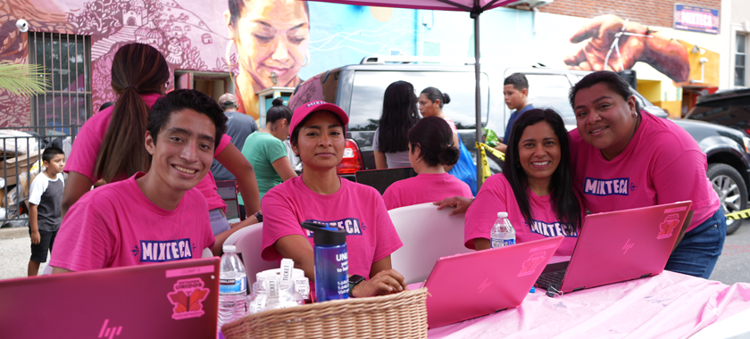 Four staff and volunteers in bright pink shirts and in front of laptops at a community event