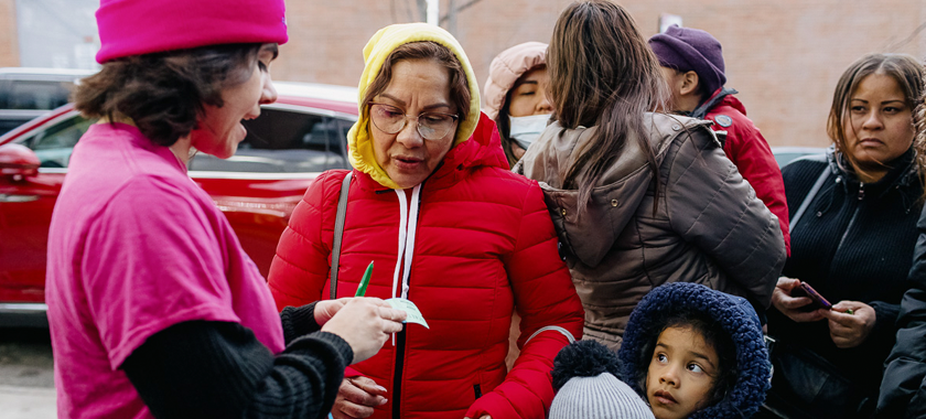 A Mixteca staff member wearing a bright pink hat and shirt speaks with asylum seekers