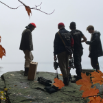 Four individuals with film and sound recording equipment stand on a boulder overlooking water and foggy terrain, framed by orange leaves