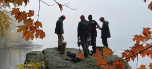 Four individuals with film and sound recording equipment stand on a boulder overlooking water and foggy terrain, framed by orange leaves