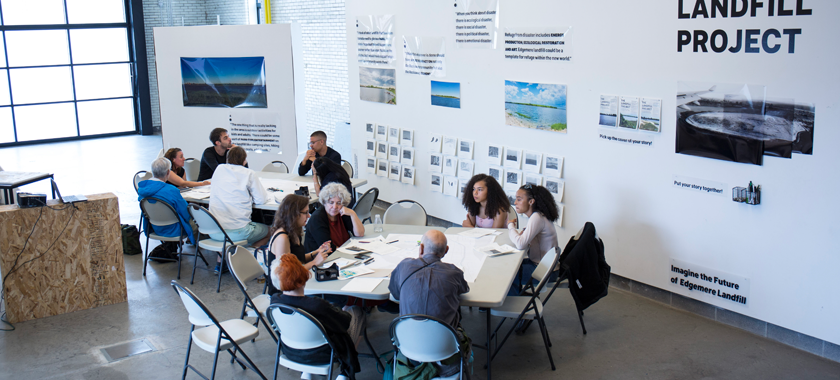 A group of people is seated at two tables surrounded by The Landfill Project exhibition featuring facts and photographs of the landfill. They have maps and drawing tools and are brainstorming ideas for the future of the site.