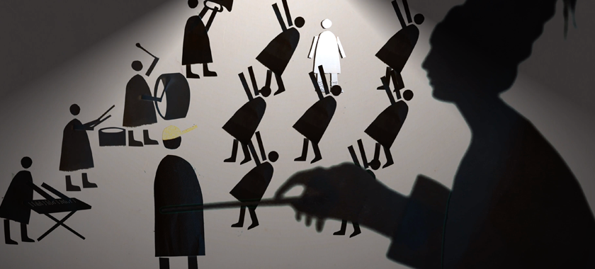 Shadowy papercraft figurines dance and play instruments while a large blurred profile of a woman dominates the scene.