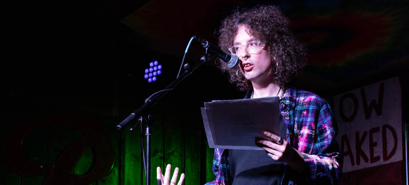 imogen, wearing bleach-purple flannel, black top & leather pants, gestures while reading onstage at Blue Moon.