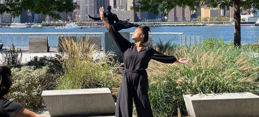 A dancer raises their arms towards the sky, in conversation with a sculpture behind them on the Queens, NY waterfront.
