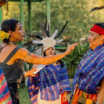 People dressed in traditional Aztec clothing stand in a circle outdoors. A woman holding a feather blesses another woman on her nose.