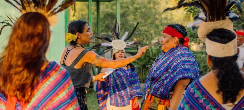 People dressed in traditional Aztec clothing stand in a circle outdoors. A woman holding a feather blesses another woman on her nose.