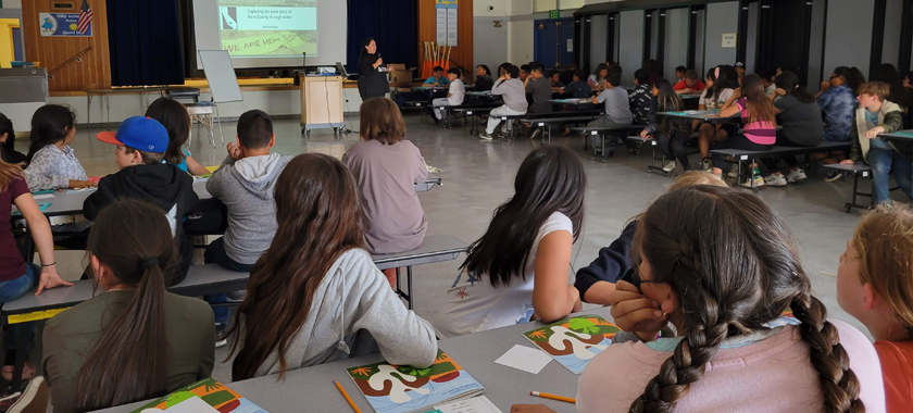 Art workshop in school cafeteria with about 90 fifth grade students sitting on tables listening to a presentation by the artist who is standing in front of large screen with projected images.