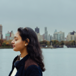 A young woman faces left with her eyes closed. Behind her is the East River and the skyline of Manhattan and Roosevelt Island. Though it's a cloudy day, birds can be seen flying behind her.