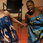 Two dancers, holding hands, moving to the side, one dance-head facing sideways, the other dancer has a huge smile on her face facing forward. Both wearing colorful african skirts. A black chair is next to the dancer on the left.