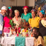 There are ten people around a festive colorful Kumina table that has colorful candles, a basket with fruit offerings, a bright colored croton plant among fine crystals and glasswares. Around the table are five female dancers and five male musicians, two of which are seated on top of their Kumina Drums. Everyone looks festive and bright.