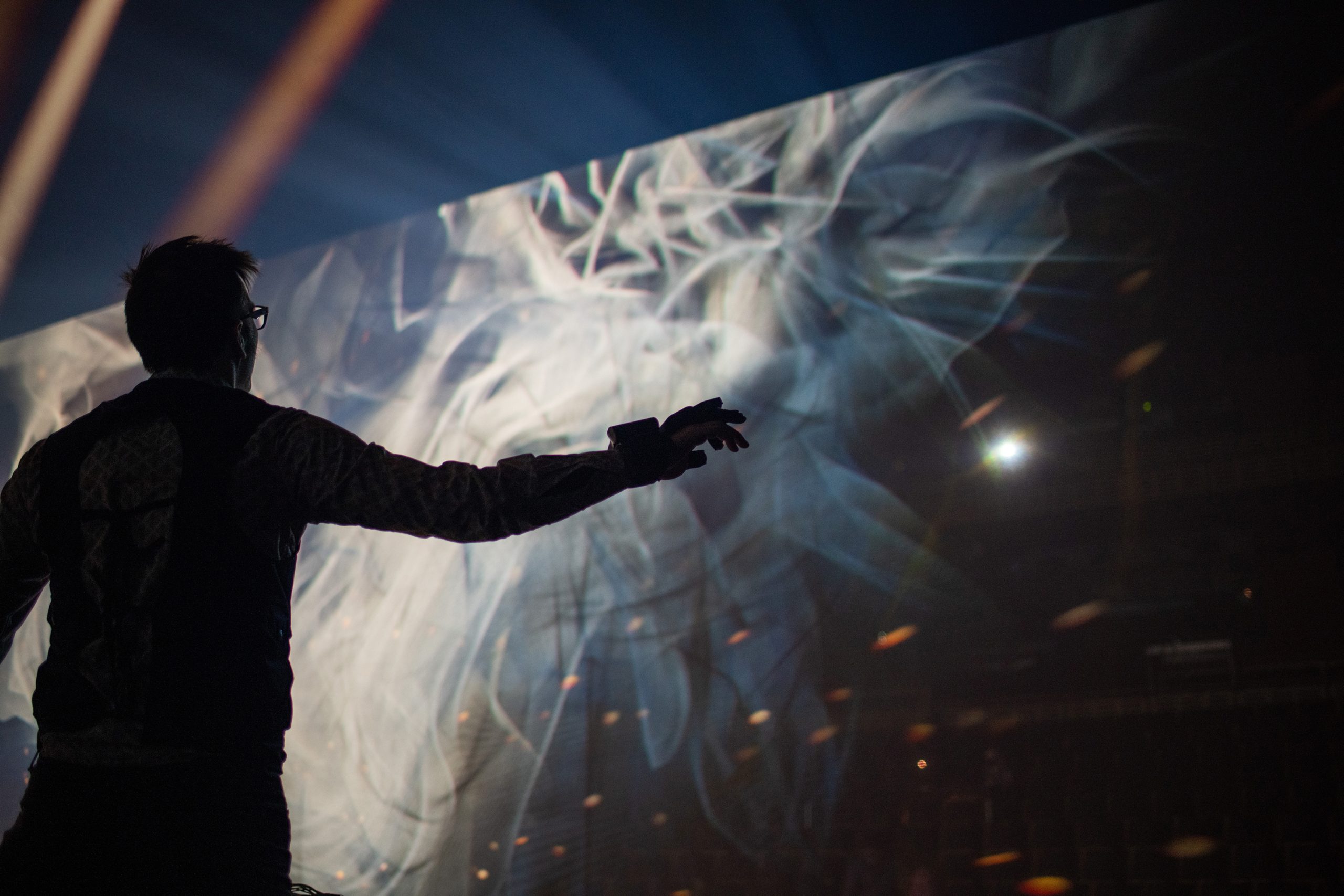 Alt Text: Male performer with gesture sensor glove is drawing graphics in the air, and the resulting scrim of projected graphics is shown, behind the figure, within the blackbox theater.