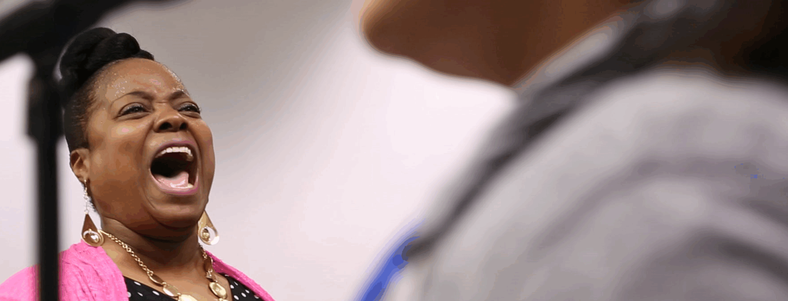 A Black women wearing a pink vest with a polka dress sings. On the right side of the frame is someone's shoulder, wearing a grey sweater.