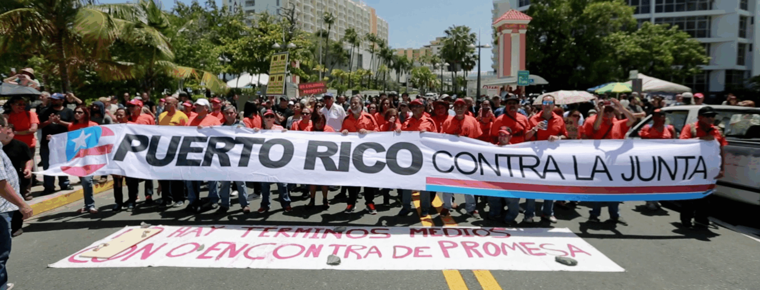 Masses of protestors hold a giant banner, "Puerto Rico Contra La Junta," (Puerto Rico against the Board) at a street protest in 2017 in San Juan, Puerto Rico