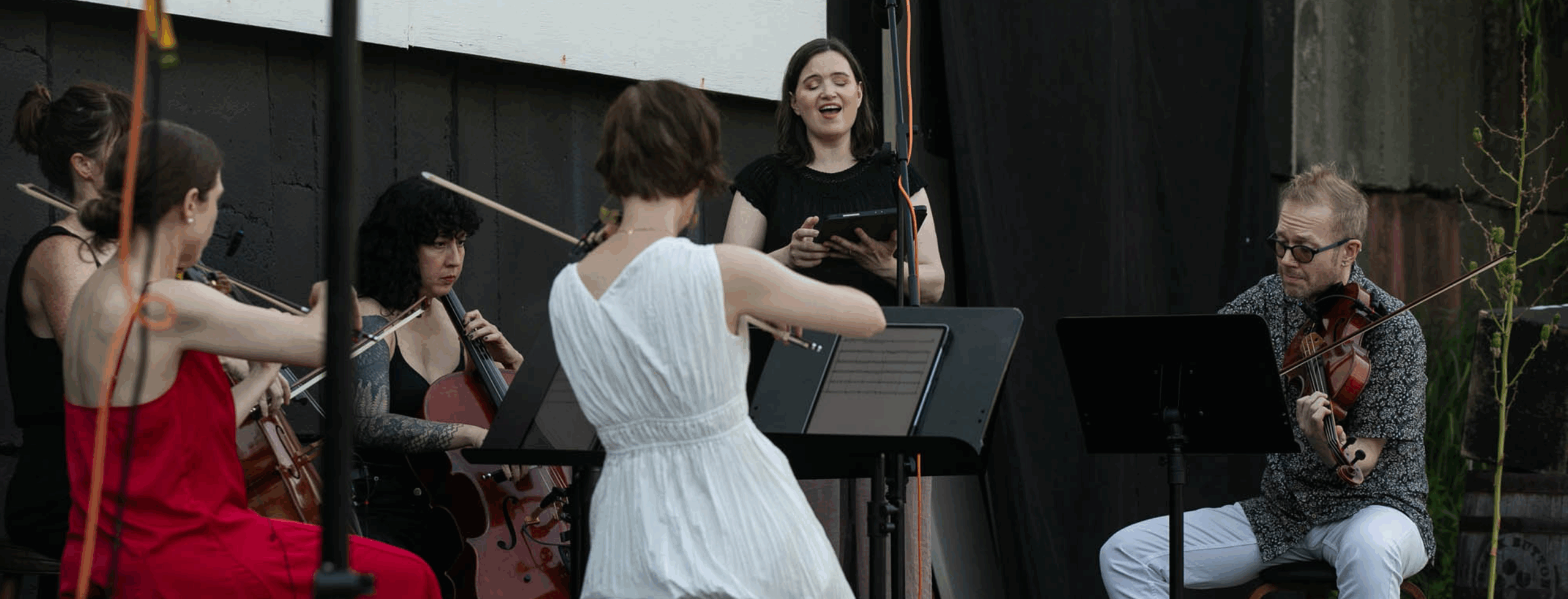 Two violinists, two cellists, one soprano, and one violist are performing Michael Nyman’s piece “No Time in Eternity” outside under a movie screen.