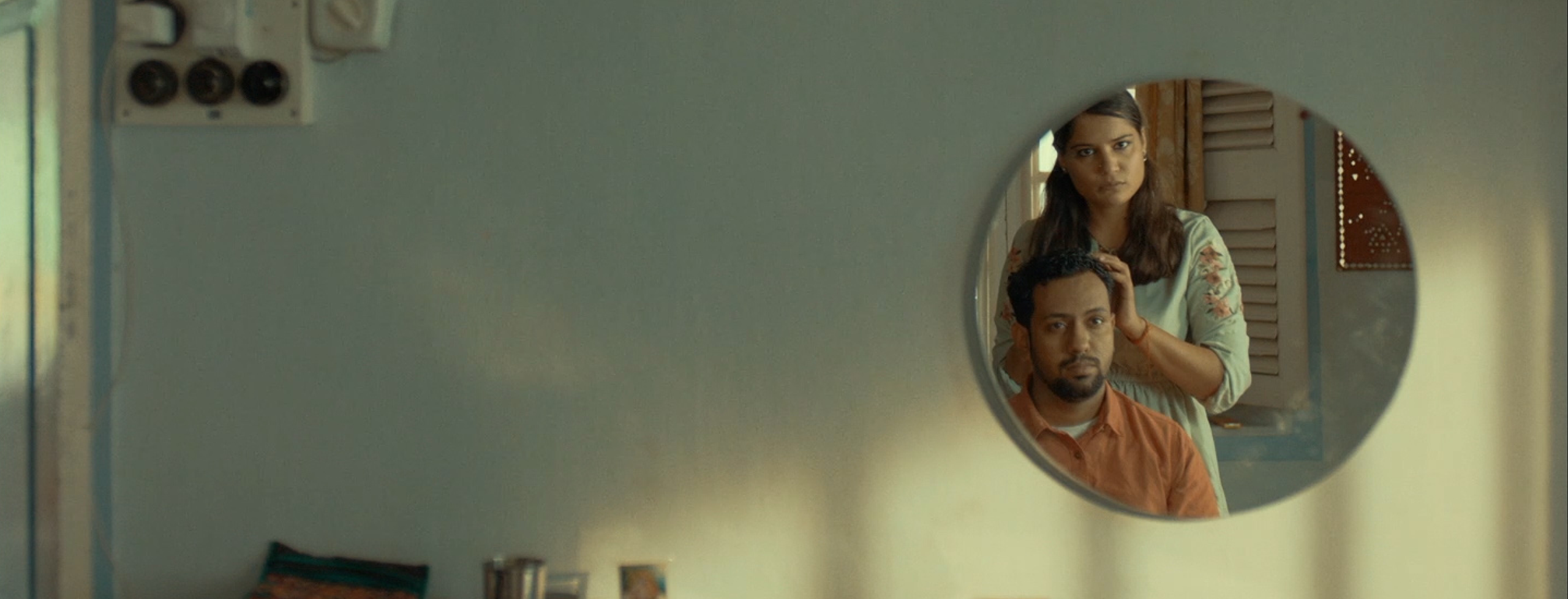 A young woman in a light green dress stands with her hands on her husband's head who is seated, dressed in an orange shirt; both staring their reflection in a round mirror on the wall.